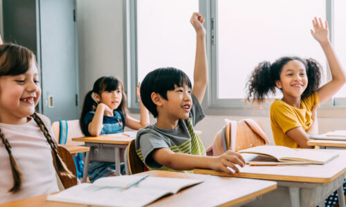 Smiling students all raise their hands while sitting at desks