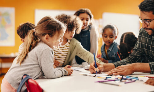 Three preschool age children watch a man draw on a child sized table