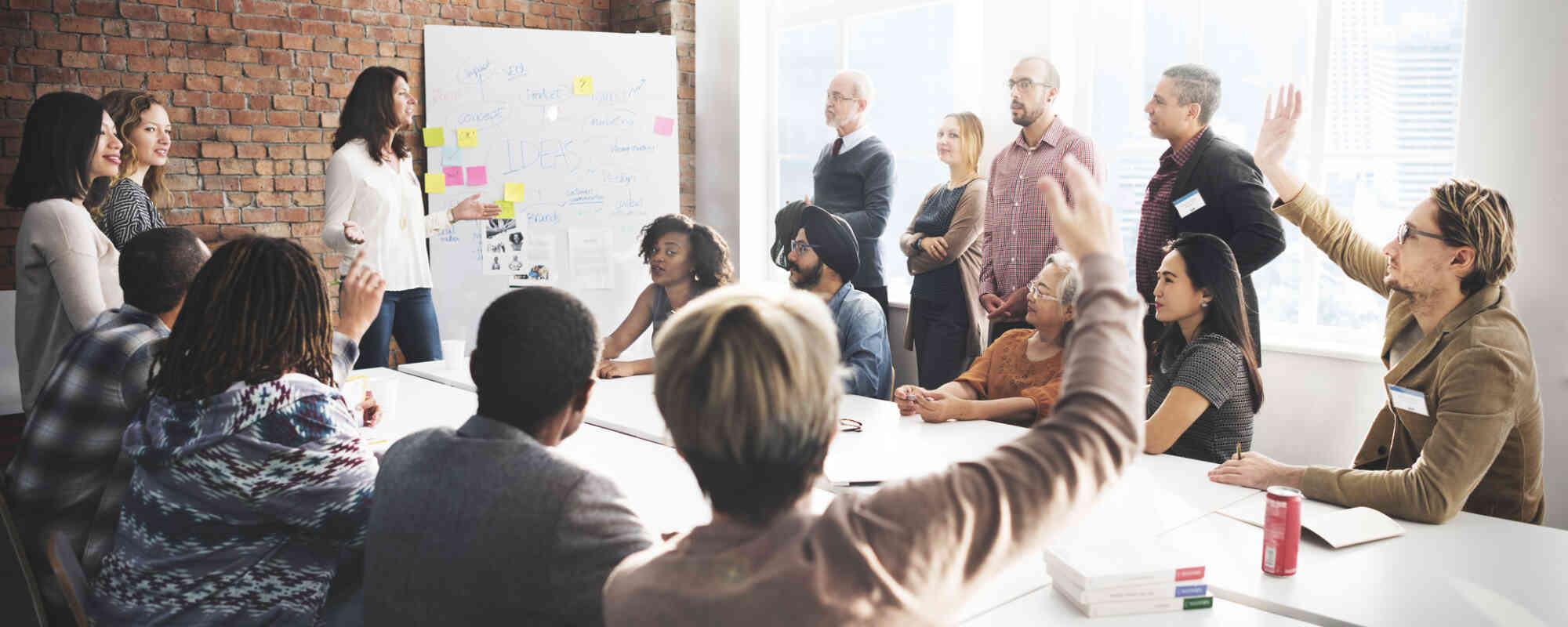 A large group of working professionals congregate around a conference table. A few are raising hands. The group is in discussion around a whiteboard with sticky notes on it.