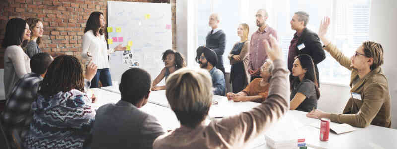 A large group of working professionals congregate around a conference table. A few are raising hands. The group is in discussion around a whiteboard with sticky notes on it.