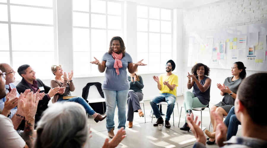 A woman stands in the center of a circle of seated people while they clap