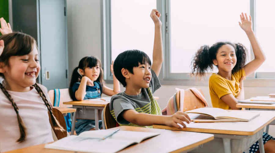 Smiling students all raise their hands while sitting at desks