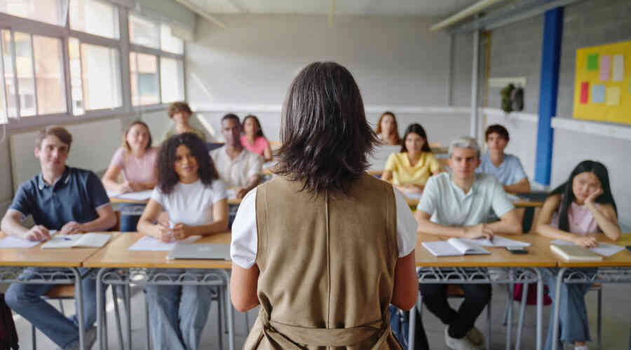 A classroom full of students face a teacher, with the camera facing the back of the teachers head