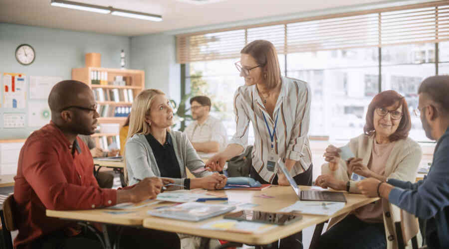 A woman leans over a table in conversation with four seated people, books and laptops are spread across the table, this takes place in a classroom