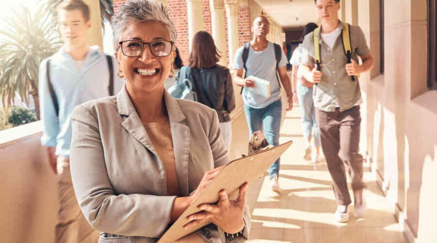 Image of a gray-haired woman, smiling at the camera, who appears to be principal in a school. Behind her, students walk through halls with their backpacks.