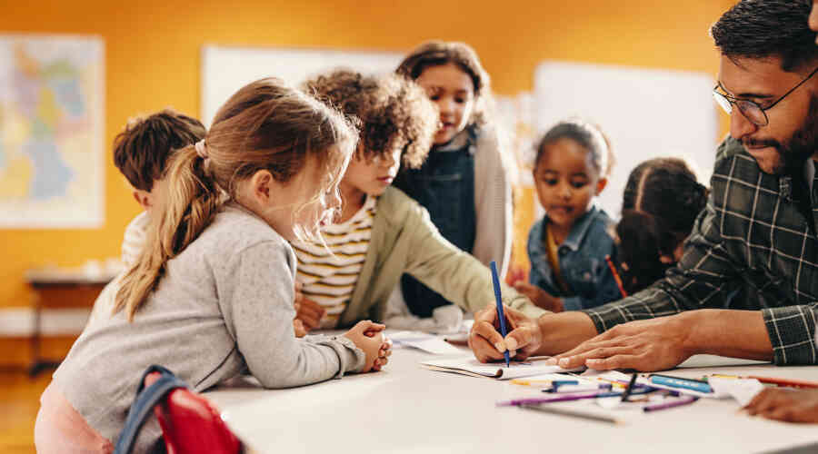 Three preschool age children watch a man draw on a child sized table