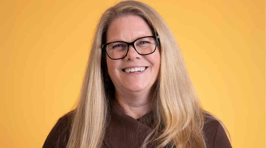 A headshot photo of Deanna Niebuhr, wearing a brown sweater, smiling against a bright yellow backdrop.