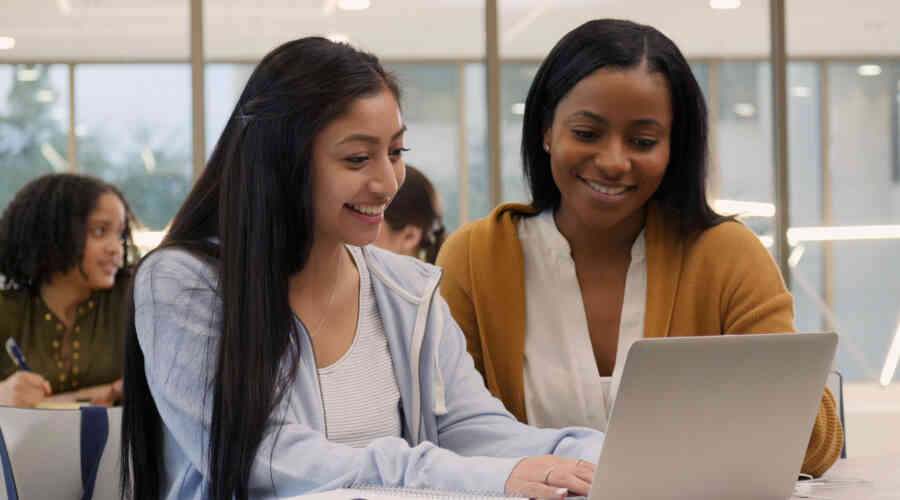 Two young women of color sit and work in front of a laptop, smiling.