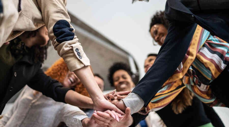 A number of young adults stand in a circle, smiling, with each of their hands in the center, stacked.