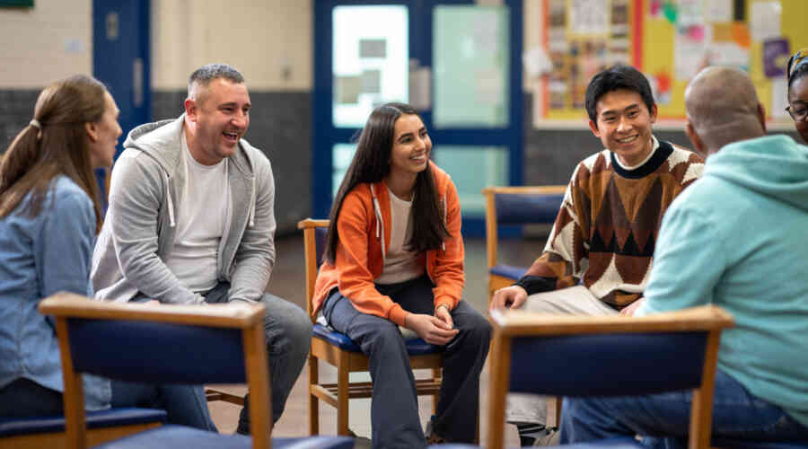 A group of students and adults sit around in a circle in a meeting, laughing, in a school setting.