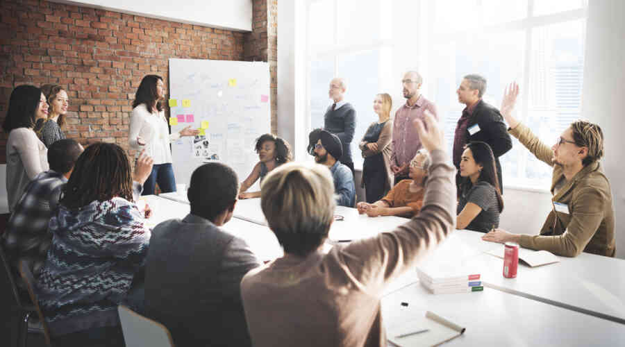 A large group of working professionals congregate around a conference table. A few are raising hands. The group is in discussion around a whiteboard with sticky notes on it.