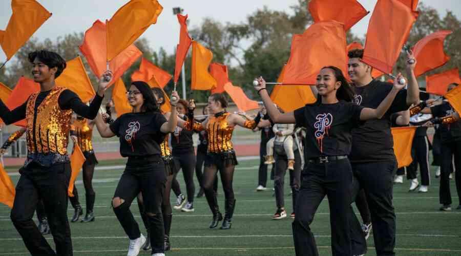 High School students march a football field with their instruments and orange flags, smiling wide.