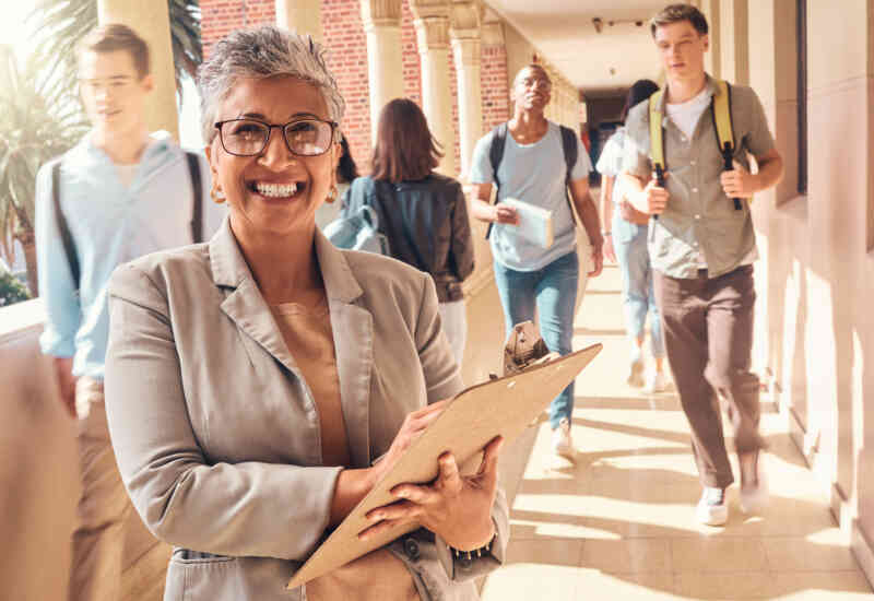 Image of a gray-haired woman, smiling at the camera, who appears to be principal in a school. Behind her, students walk through halls with their backpacks.