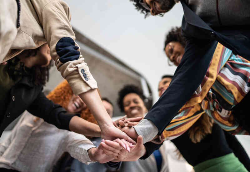 A number of young adults stand in a circle, smiling, with each of their hands in the center, stacked.