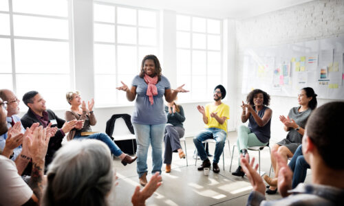 A woman stands in the center of a circle of seated people while they clap