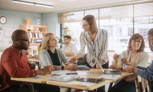 A woman leans over a table in conversation with four seated people, books and laptops are spread across the table, this takes place in a classroom