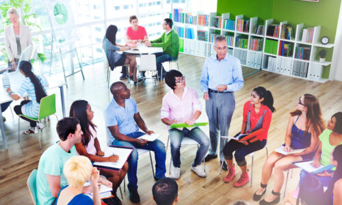 A group of young people sit in a circle on wireframe chairs listening to an older speaker, in what appears to be a classroom or other learning space. In the background, two small groups appear to be learning too