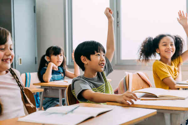 Smiling students all raise their hands while sitting at desks